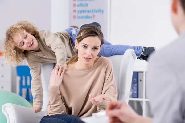 Mère fatiguée qui discute avec un professionnel pendant que ses enfants énergiques grimpent sur le canapé derrière elle