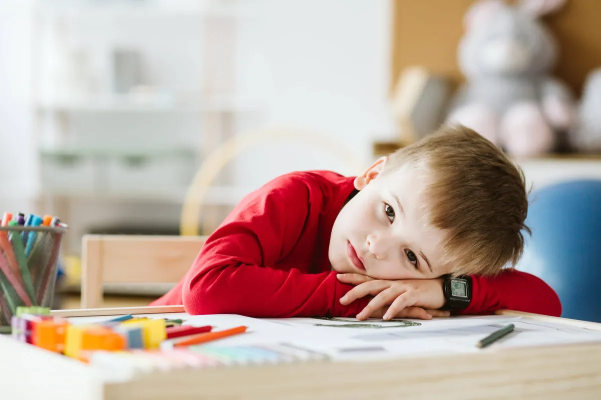 Garçon qui s'ennuie, la tête appuyée sur ses bras à un bureau couvert de crayons de couleur