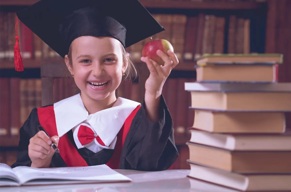 Fillette souriante en toge et mortier de finissante, une pomme à la main, à côté d'une pile de livres