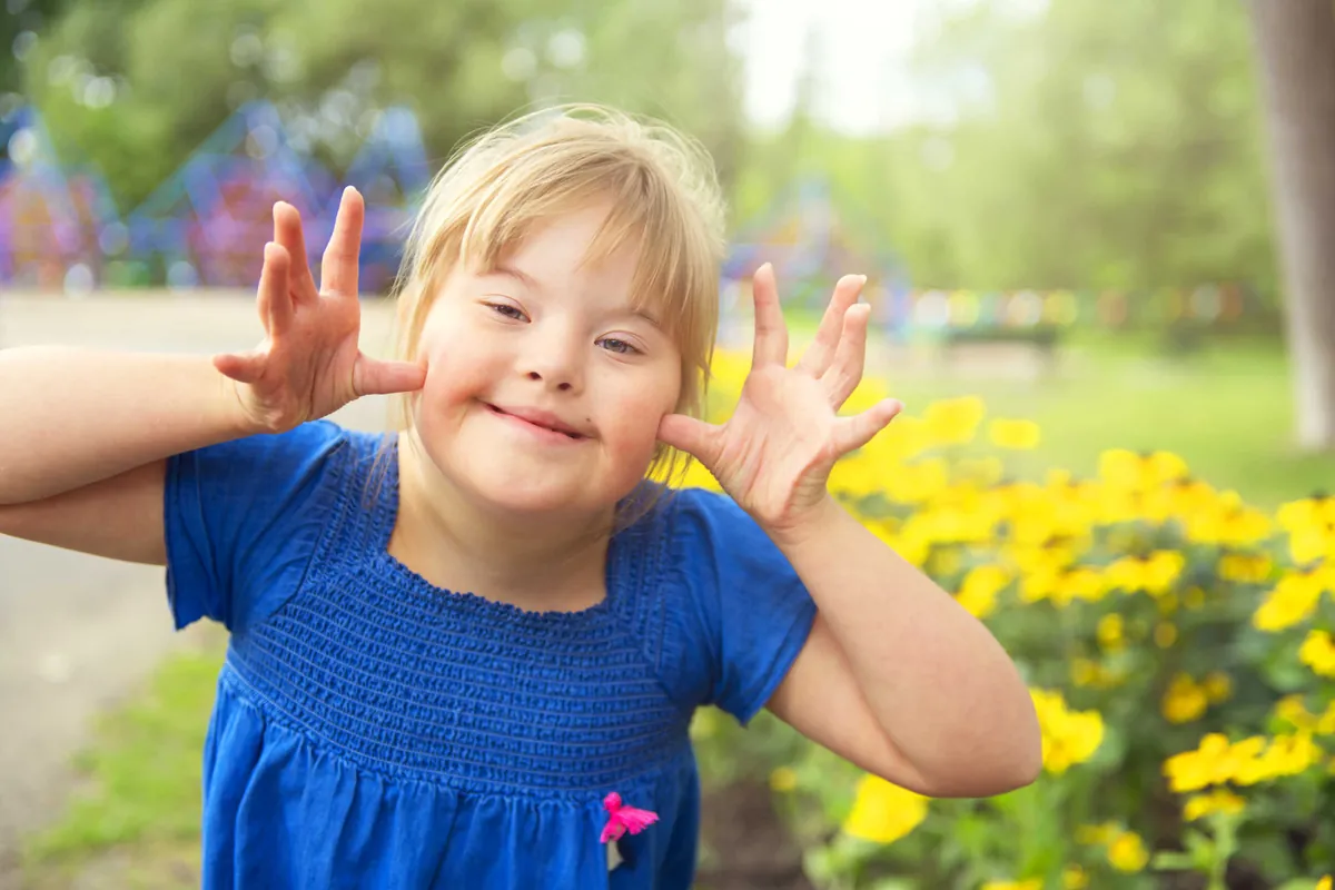 Fillette souriante ayant une trisomie 21 qui fait une grimace enjouée dans un jardin de fleurs jaunes