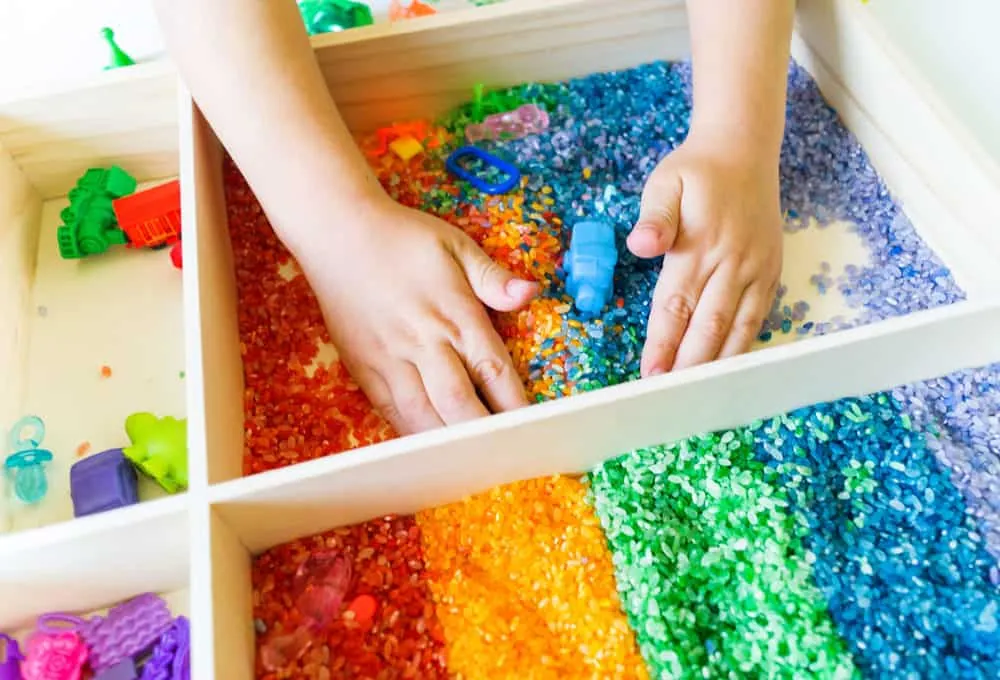 Mains d'enfant plongées dans un plateau sensoriel de riz aux couleurs de l'arc-en-ciel