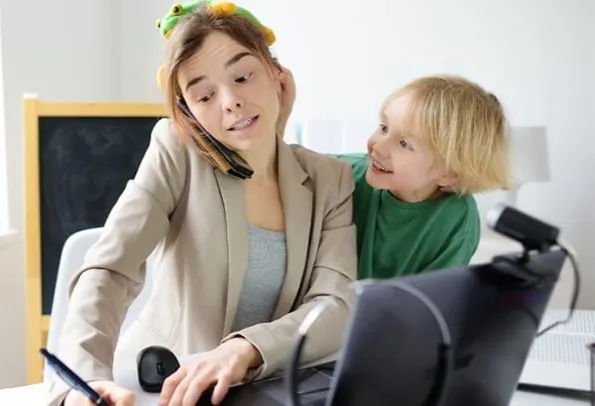 Parent occupée qui travaille à l'ordinateur et au téléphone pendant que son jeune garçon s'accroche à ses épaules