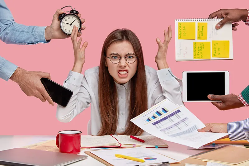 Jeune fille dépassée, entourée de téléphones, de papiers et d'un réveil, illustrant la surcharge de tâches