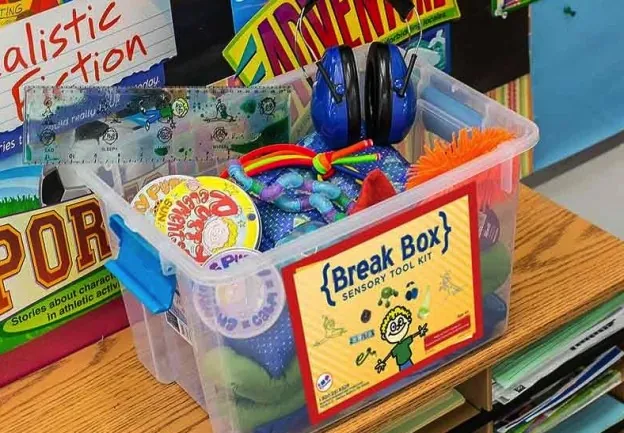 Bac de plastique transparent étiqueté « Break Box » contenant des fidgets, des balles antistress et des coquilles antibruit sur un pupitre