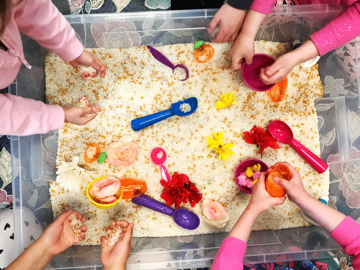 Mains d'enfants qui puisent du riz, des fleurs et de petits objets dans un bac sensoriel thématique