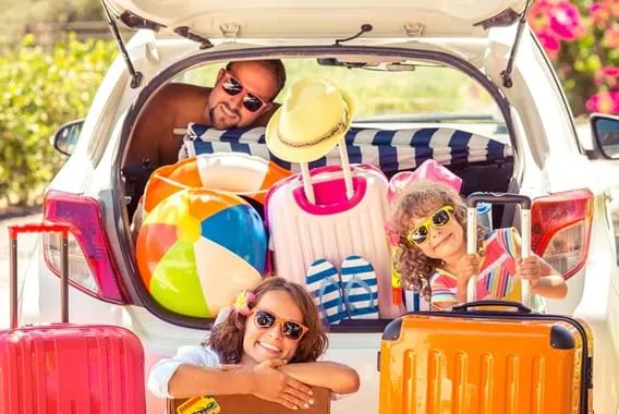 Famille en lunettes de soleil qui pose dans le coffre d'une voiture rempli de valises, d'un ballon de plage et d'un chapeau