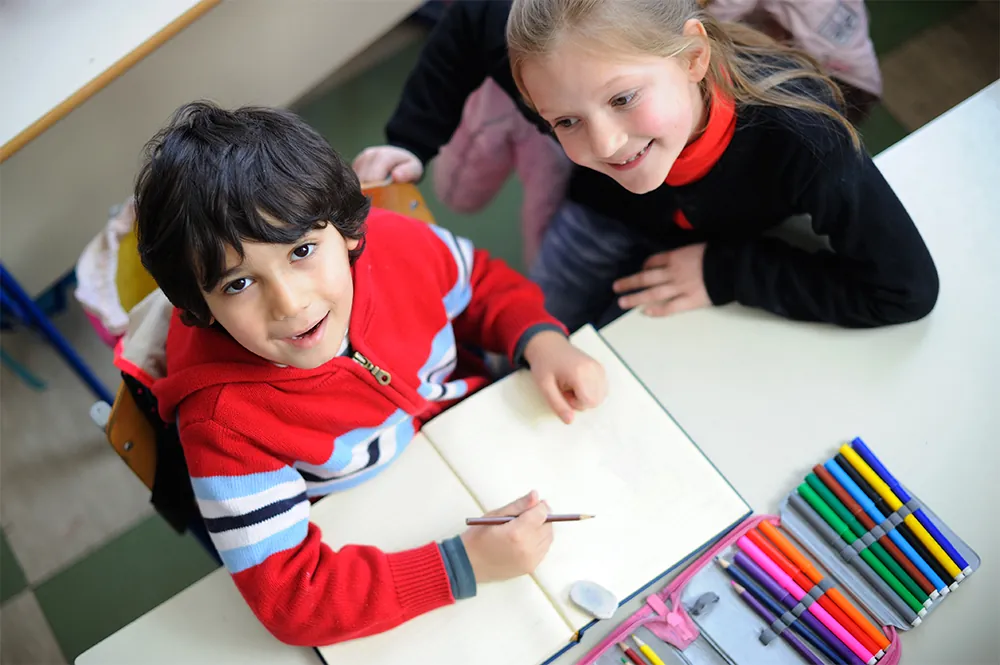 Deux écoliers à un pupitre avec des crayons feutres et un cahier, l'un regardant l'objectif